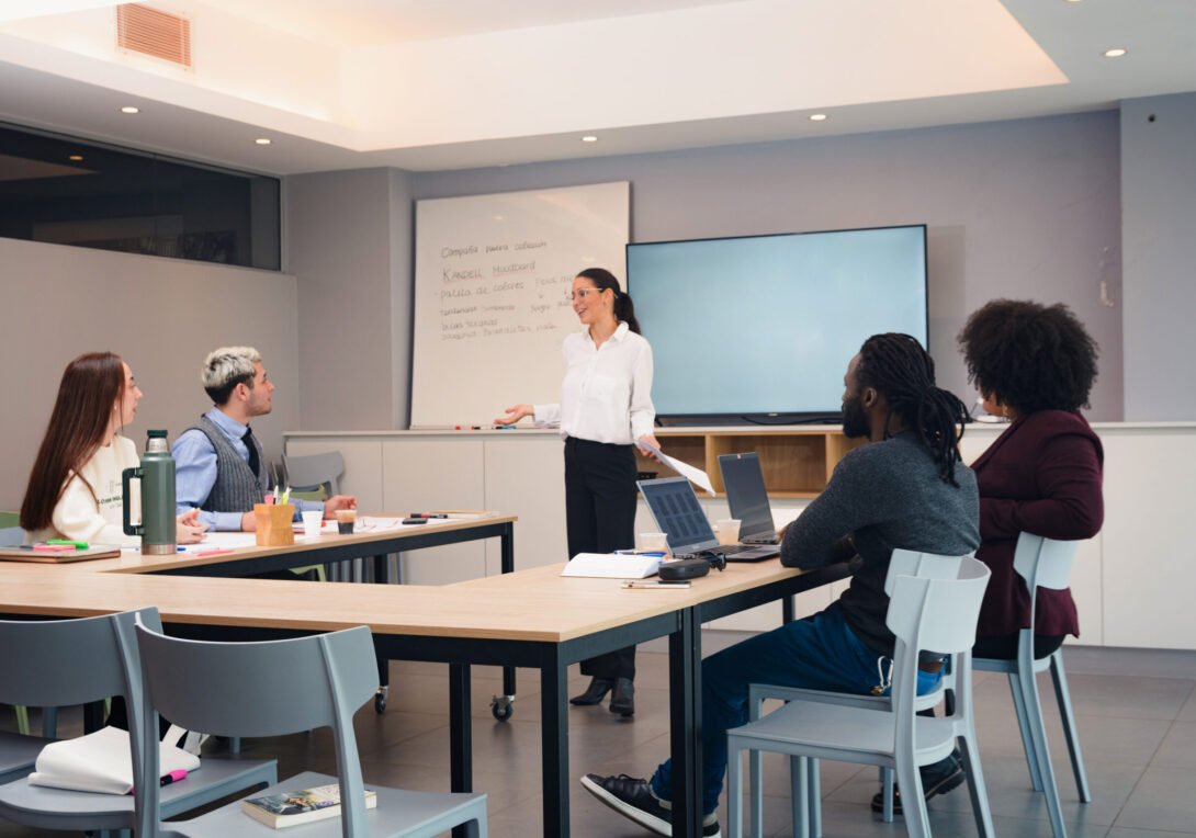 Woman standing in the middle of the room ending the meeting in the office, she has papers in her hands and is gesturing with her body while she speaks, there is a blank screen behind her.